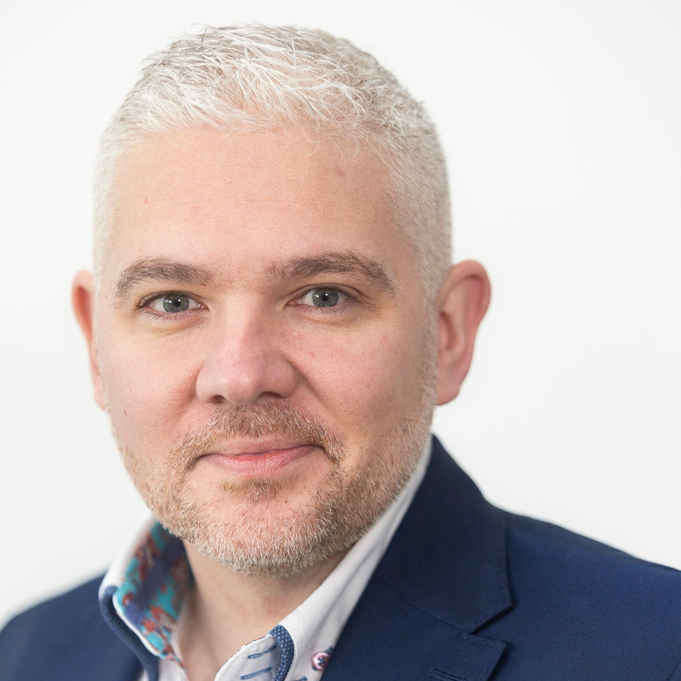 Male entrepreneur with grey hair and beard wearing a blue suit and patterned shirt on a white background