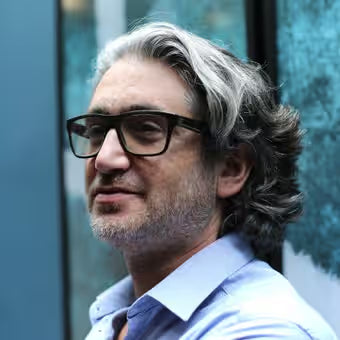 Male entrepreneur with glasses and grey hair wearing a light blue shirt against a blurred outdoor background